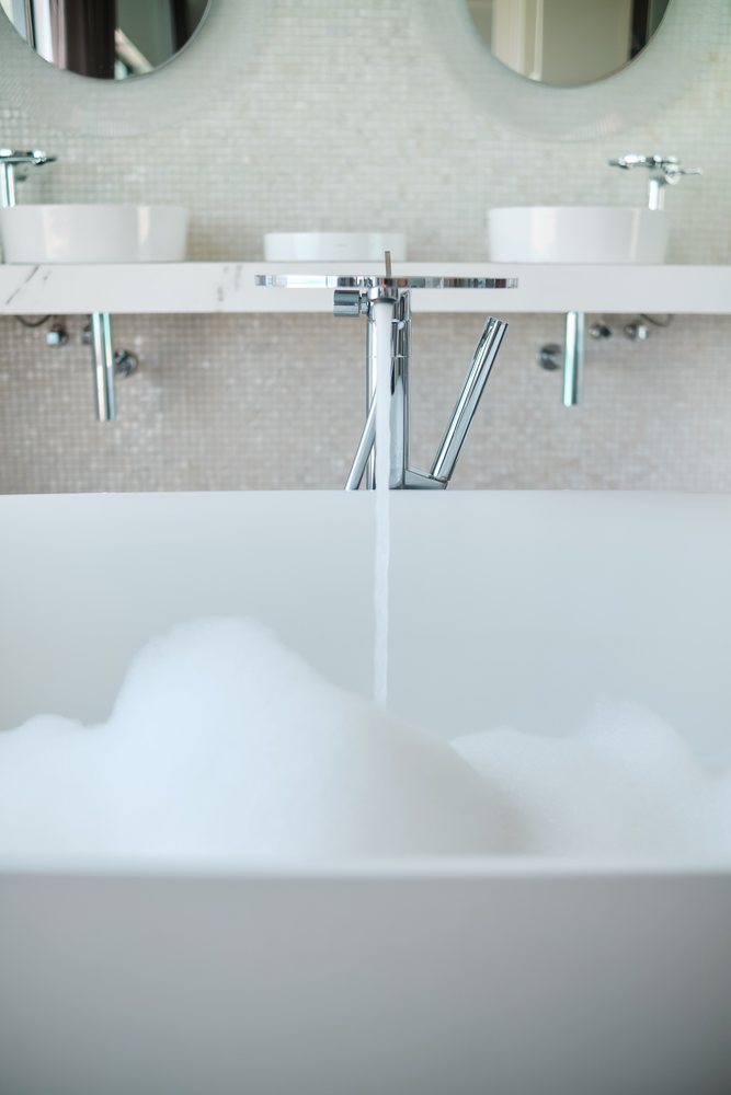 A modern white bathtub in a well-lit bathroom filling with foamy bath water from a running tap, Maldives.