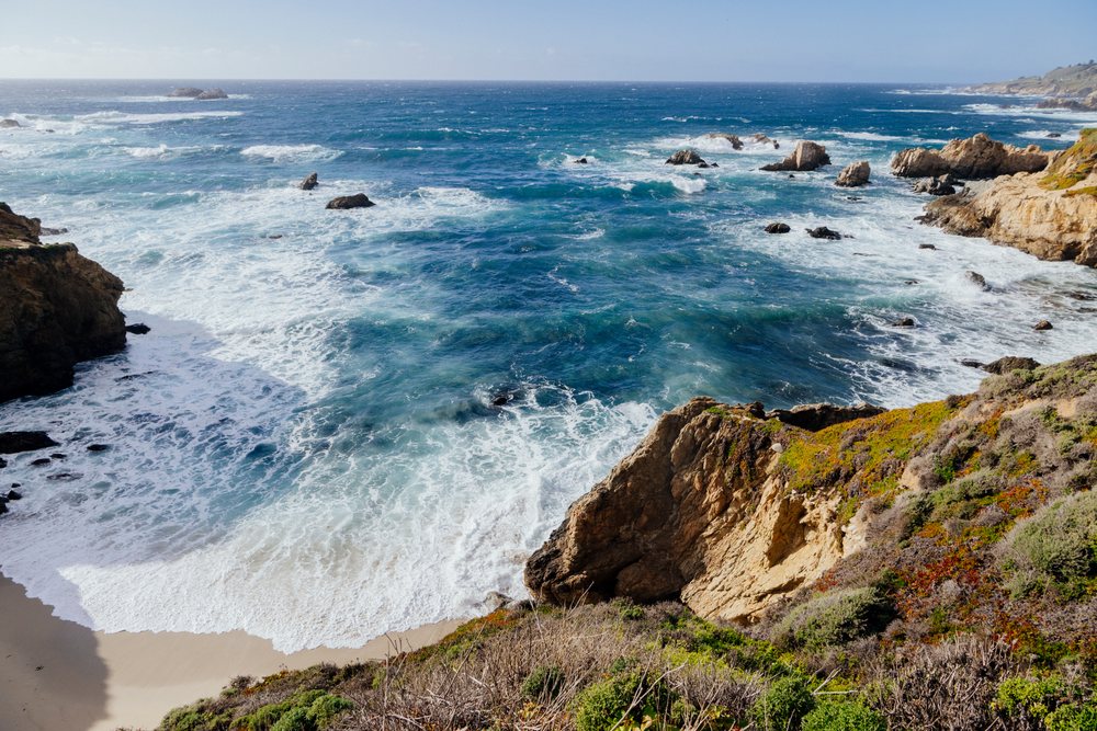 Ocean waves crashing along a rocky coastline with cliffs, beach sand, and vegetation on the California coast under a clear sky, USA.