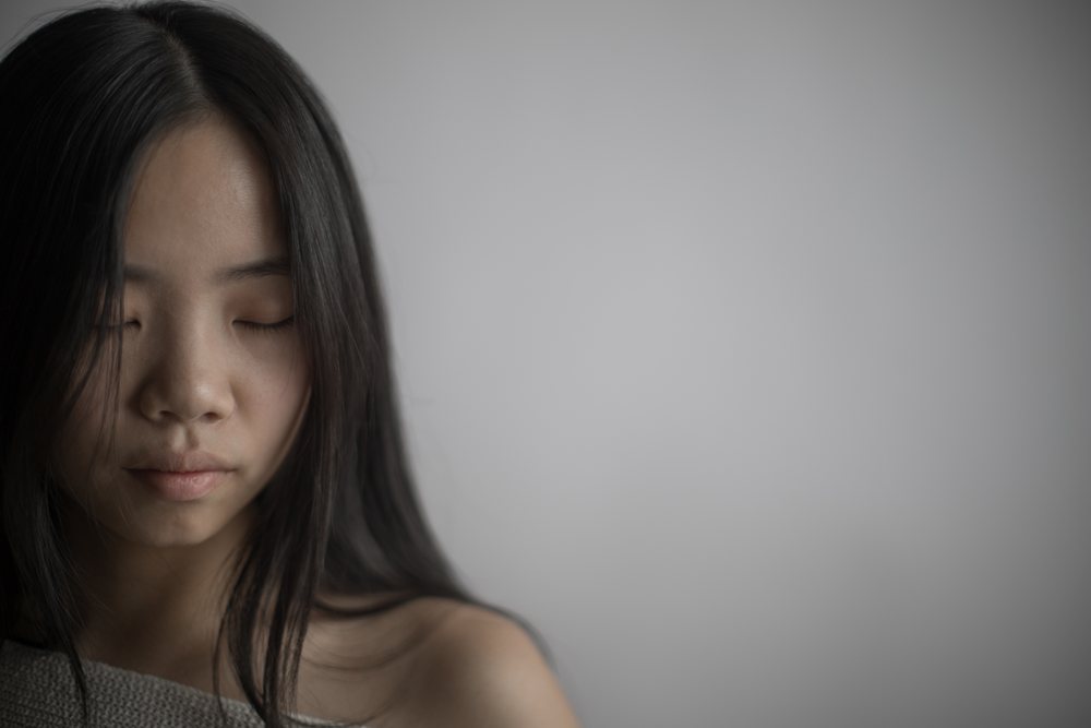 Serene portrait of a woman with closed eyes and black hair in a softly lit indoor setting against a gray background, Art, emotional.