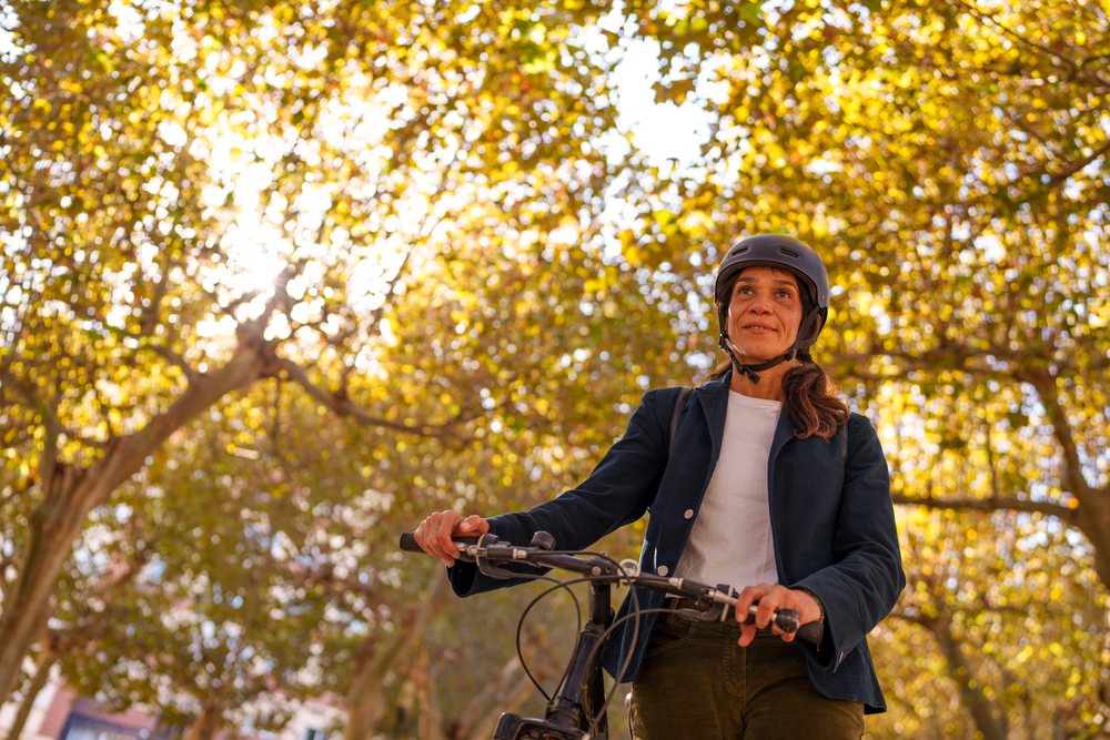 Woman wearing a helmet rides a bicycle outdoors under autumn trees with sunlight filtering through the leaves, Málaga.