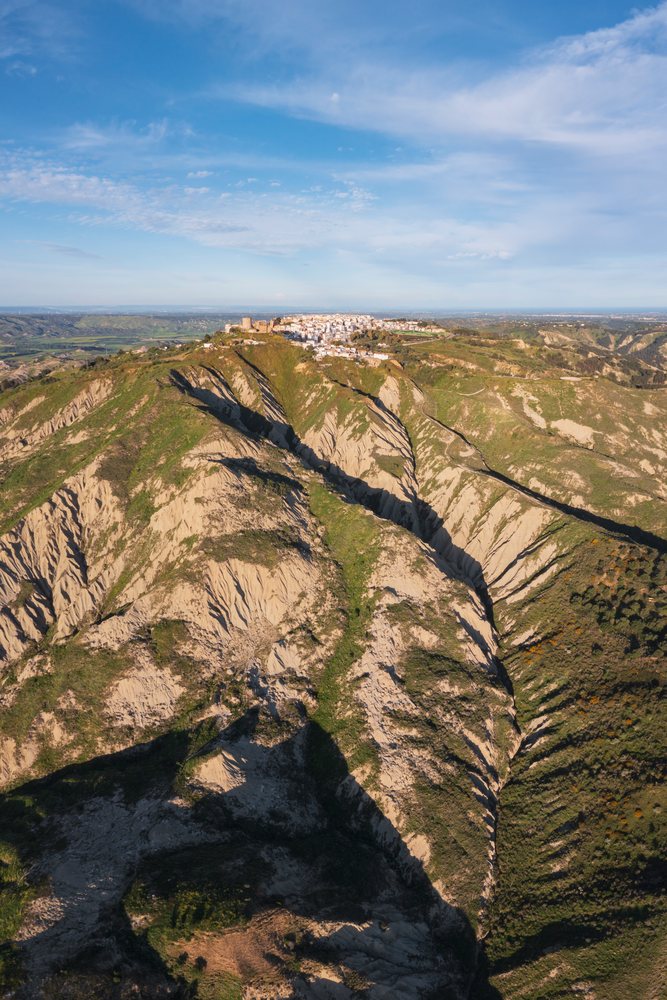 Hill town of Pisticci atop rugged eroded Calanche hills, under a blue sky with visible valley and distant plains, Basilicata, Italy.