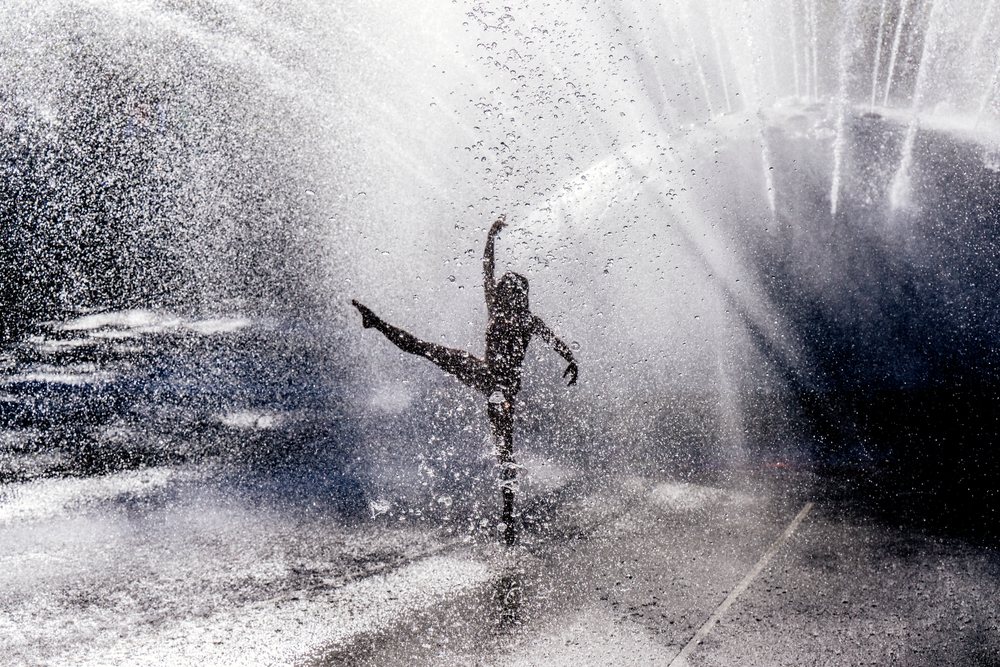 Dancer performing an acrobatic yoga pose under a fountain water spray outdoors, partner, acrobatics, USA.