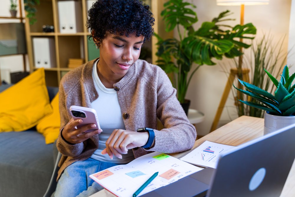 Woman sitting at a desk in a living room, checking her smartwatch while using a smartphone and planner, Spain.