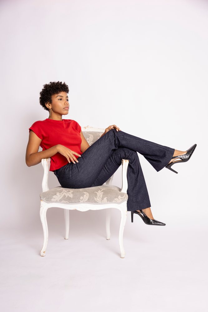 Woman with short curly hair wearing a red short sleeve top, dark jeans, and black heels poses seated on an elegant floral armchair against a white studio background, fashion, clothes, young culture.