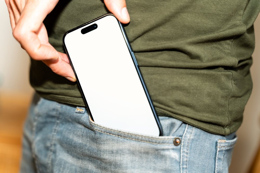 Hand holding a smartphone with a blank white screen partially inserted into a blue denim jeans pocket, with a green shirt visible, mock up, Spain.