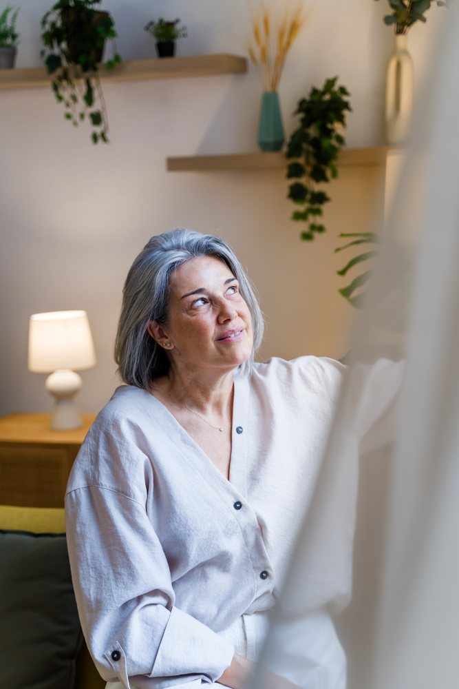 Middle-aged woman with gray hair seated on a couch, looking up inside a living room with modern decor and wooden furniture, Spain.