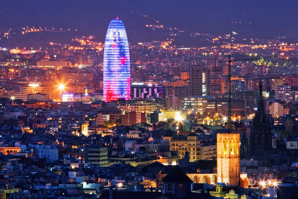 Cityscape of Barcelona at night featuring the illuminated Torre Glòries and surrounding urban buildings on a hilly background.