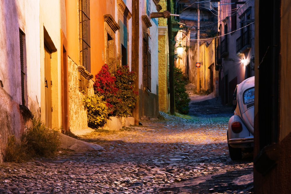 Cobblestone alley with colorful buildings, flowering shrubs, and a vintage car, Mexico, Dusk, San Miguel.