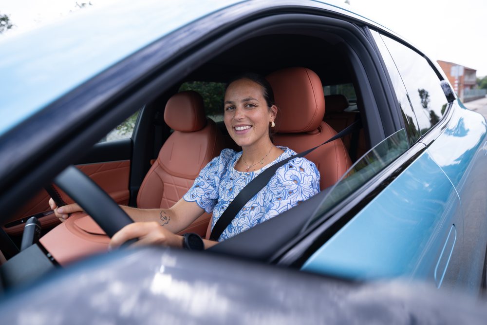 Woman smiling while driving a blue car with tan leather seats and wearing a seat belt on a road, Spain.