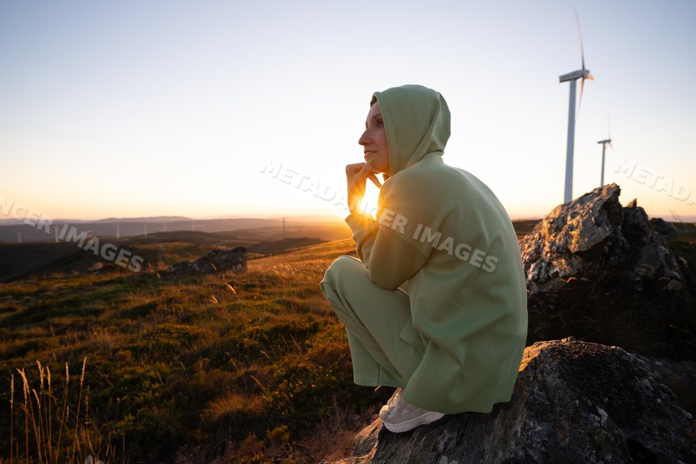 White Woman sitting on rock at sunset with wind turbines on hillside