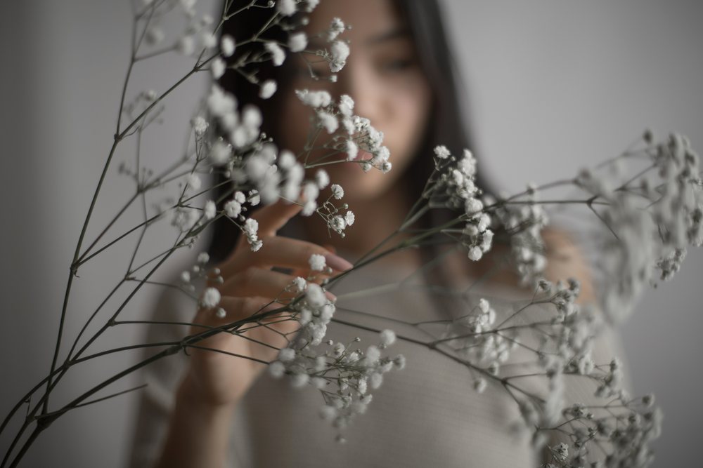 Woman gently touches delicate white baby's breath flowers in a softly lit indoor portrait with a blurred face in the background, Art, emotional.