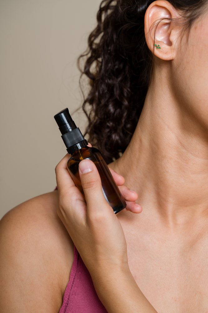 Woman with curly hair and ear piercing holds an amber spray bottle near her neck during a beauty shoot, Spain.
