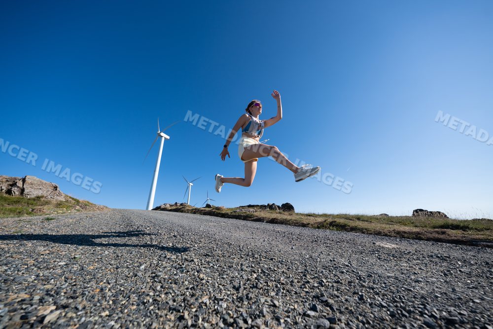 Young white woman jumps outdoors near wind turbines in Galicia, Spain