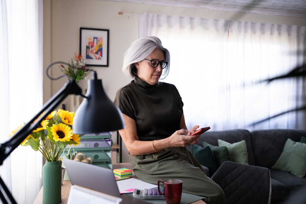 Mature woman with gray hair and glasses uses a smartphone while sitting at a desk in a modern home office living room with sunflowers and office supplies, Spain.