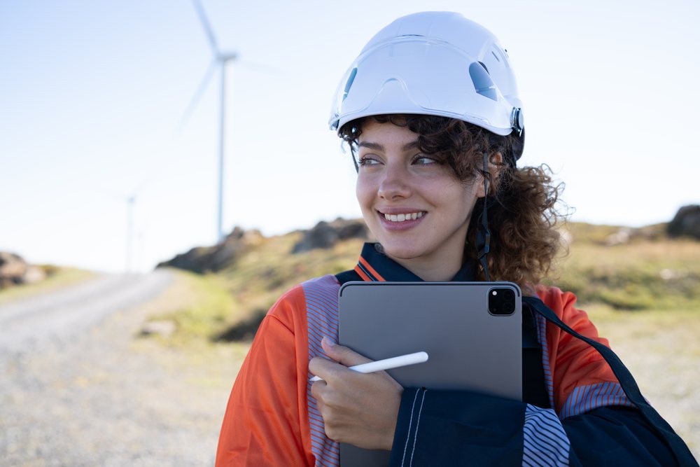 Woman engineer wearing a hard hat and orange safety jacket holds a tablet and stylus outdoors at a wind farm, sustainability, renewable energy, Galicia, Spain.