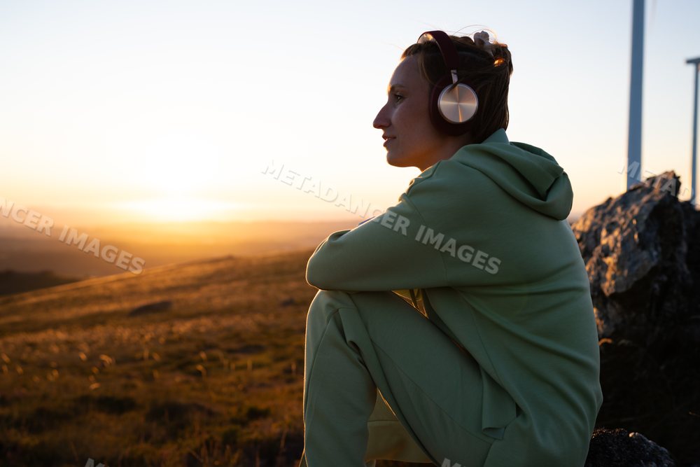 A white woman sits wearing headphones outdoors during sunset