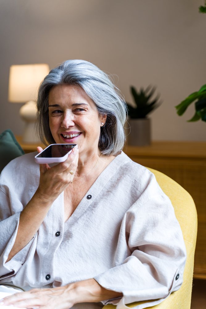 Senior woman speaking into smartphone while sitting on a yellow chair in a modern living room, Spain.
