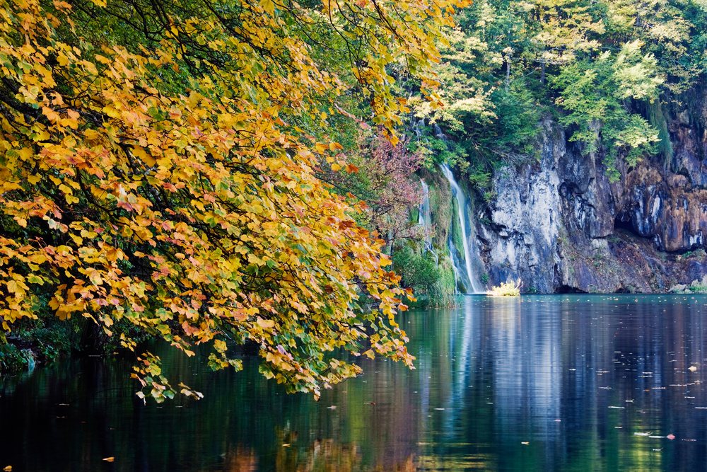 Autumn foliage with vibrant leaves beside a tranquil lake reflecting waterfalls and rocky cliffs, Croatia.