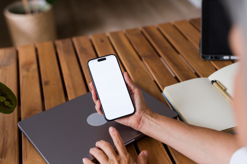 Person holding a smartphone with a blank screen over a wooden desk with a closed laptop, notebook, and pen, Spain.