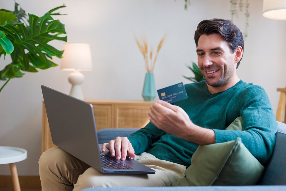 Man sitting on a couch holding a credit card and using a laptop in a cozy living room, Technology, young adult, home.