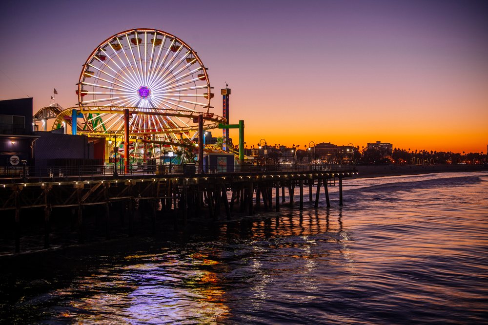 Santa Monica Pier illuminated Ferris wheel at sunset reflecting on Pacific Ocean water with skyline and palm trees visible, CA, USA.
