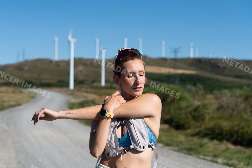 A white woman stretches her arm on a rural trail near wind turbines
