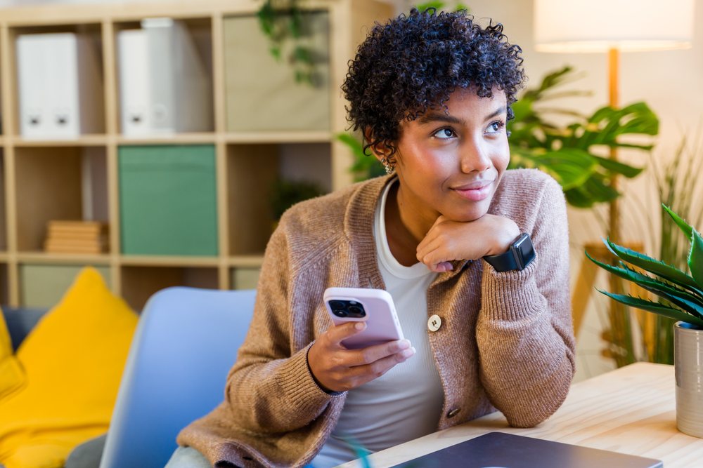 Woman with curly hair wearing casual clothing sits at desk holding smartphone, looking thoughtfully away in a modern indoor office with plants and lamp, Spain.