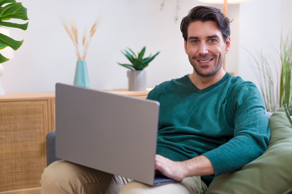 Smiling man sits on a couch using a laptop in a modern home with indoor plants and decorative vases, Technology, young adult.