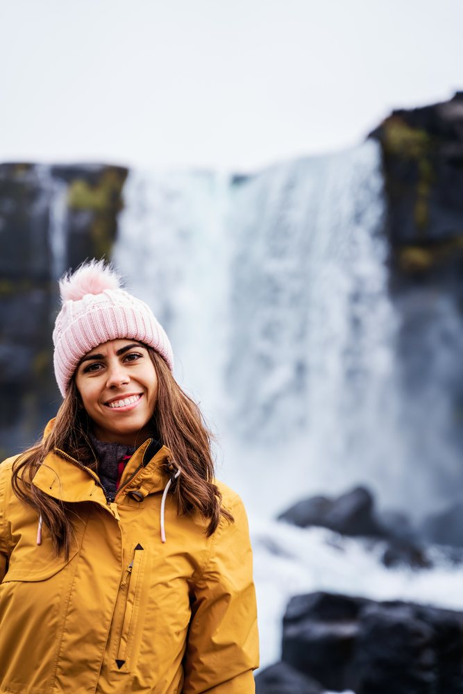 A young woman in a yellow jacket and pink knit hat smiles in front of a waterfall, Iceland.