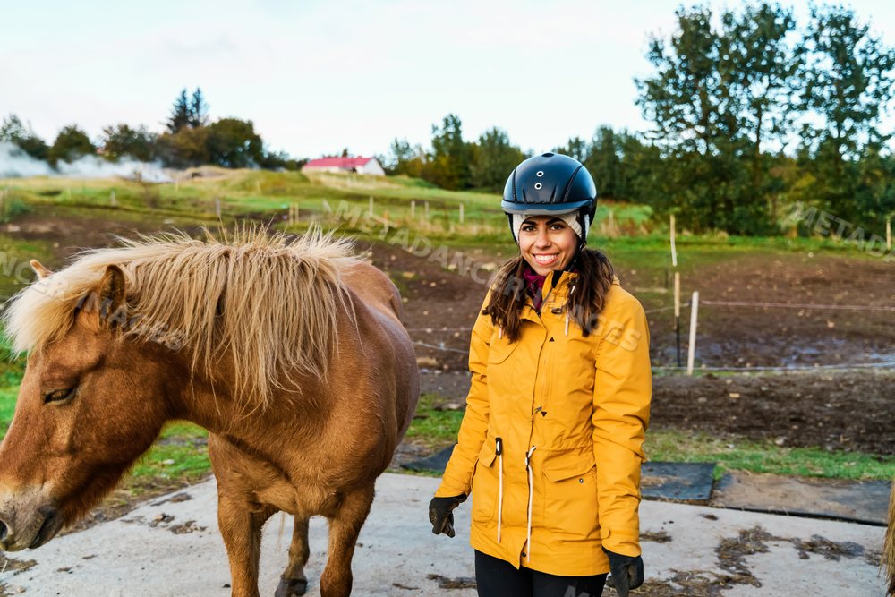A white woman stands smiling beside a brown horse on a farm
