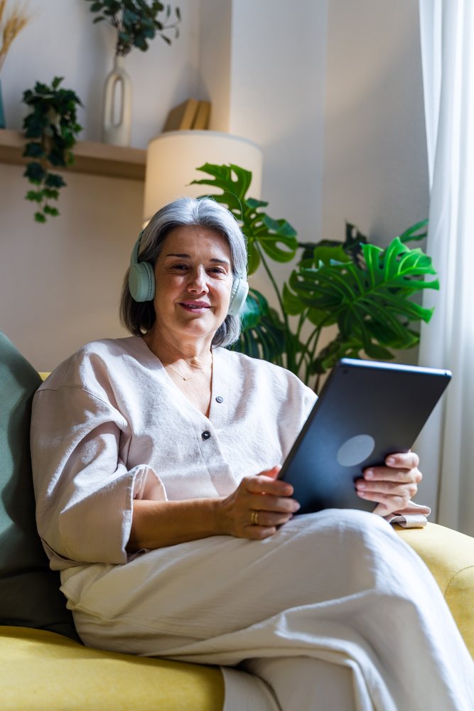 Woman with gray hair wearing headphones sits on a yellow sofa holding a tablet in a bright indoor living room with a lamp and houseplant, Spain.