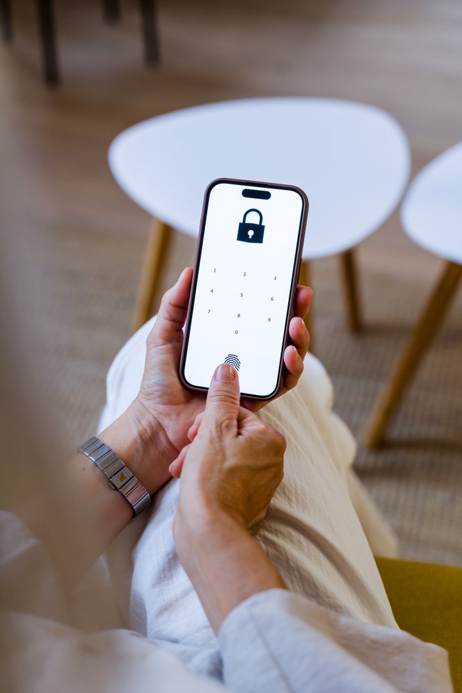 Hand holding a smartphone with fingerprint authentication and lock icon on screen indoors near white chair, Spain.