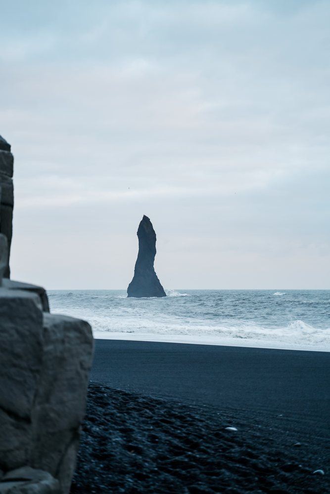 A tall sea stack rises from the ocean at a black sand beach in a beach