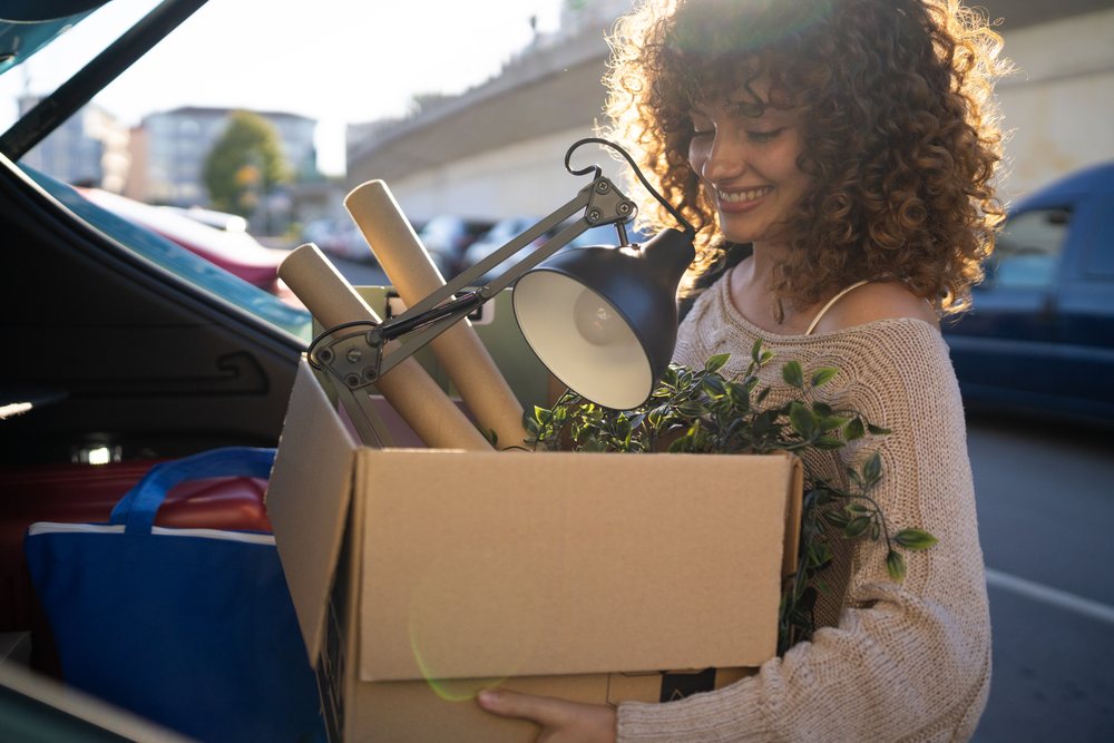 Woman with curly hair smiling while carrying a cardboard box containing a lamp, paper rolls, and a green plant outdoors in an urban setting during moving, Spain.