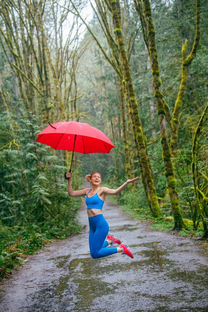 Woman in blue sportswear jumps joyfully on a wet forest trail holding a red umbrella in the rainy USA woodland.