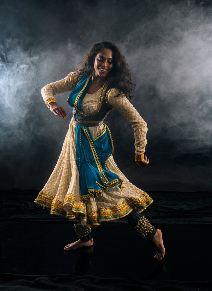 Indian classical dancer performing with ankle bells in dress and blue scarf in a dark indoor studio setting, spirituality, expression.