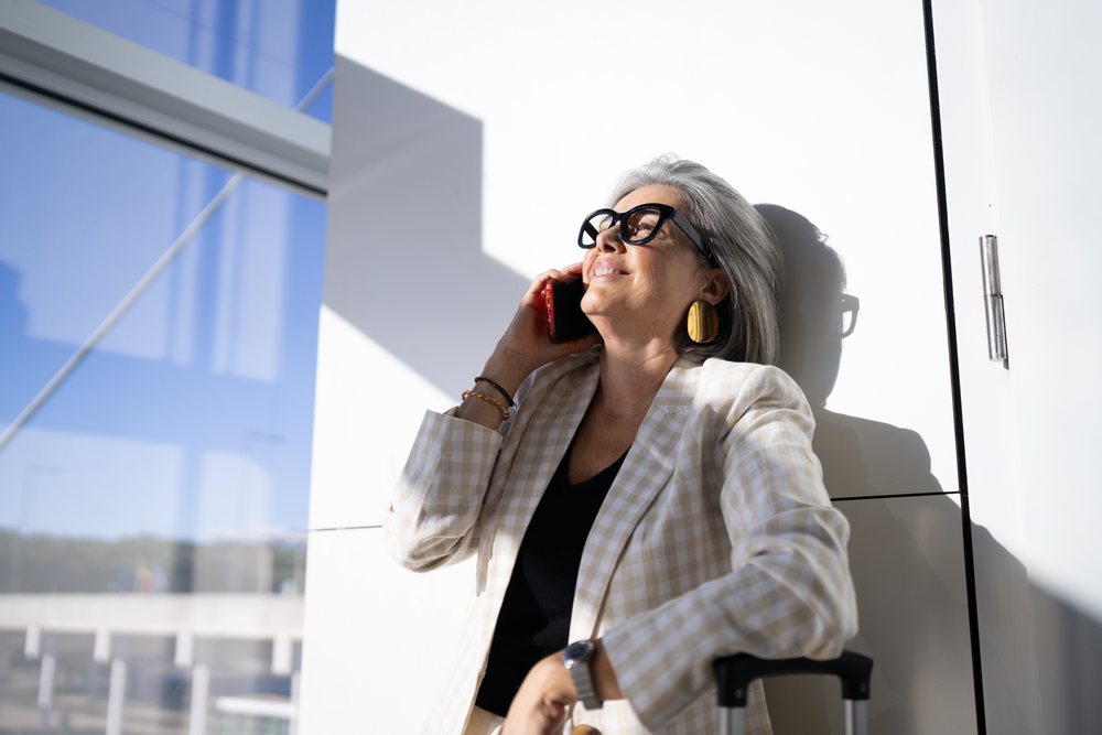 Businesswoman with gray hair and glasses making a phone call while leaning against a wall in a bright indoor setting, Spain, Madrid.