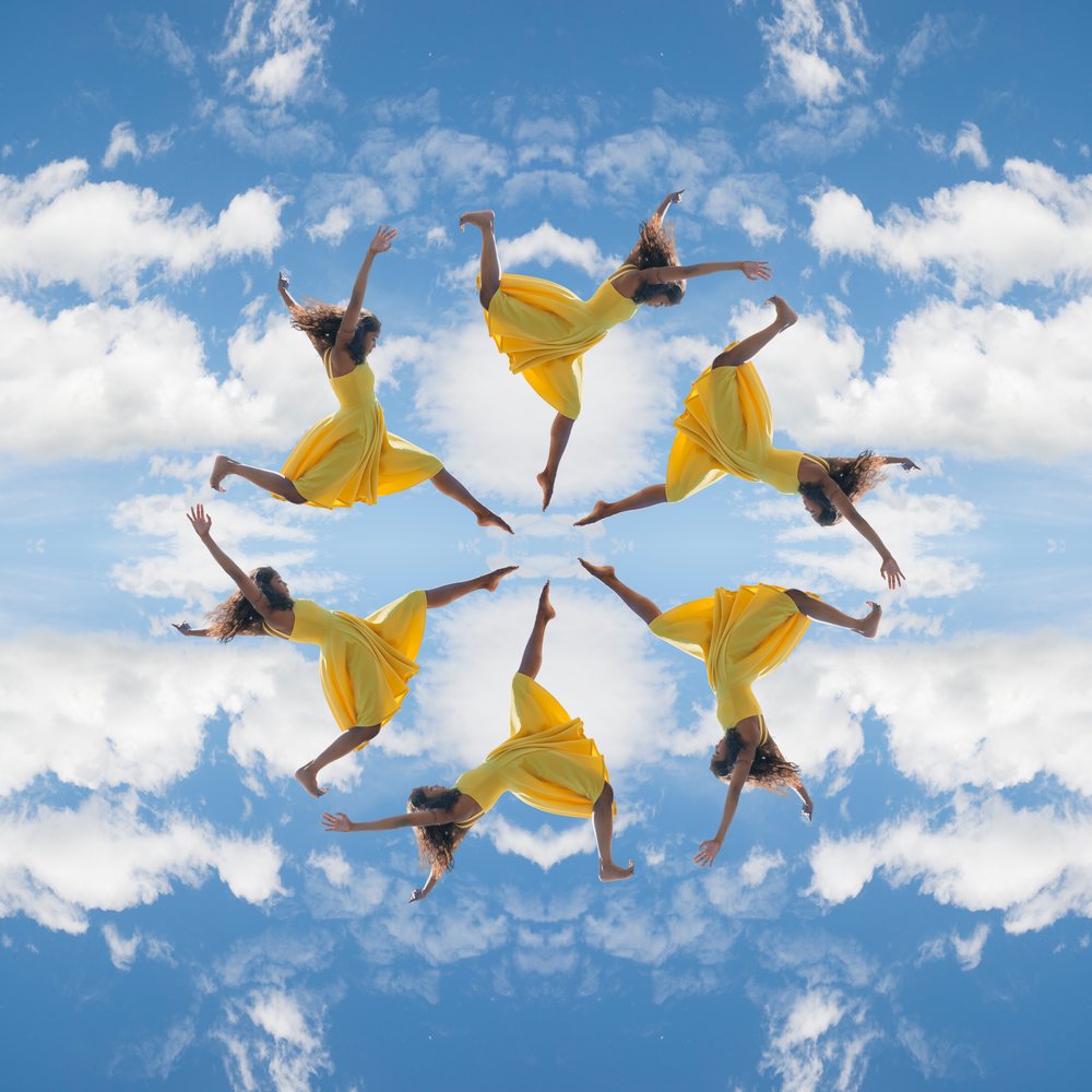 Woman in a yellow dress jumps dynamically against a blue sky with scattered clouds arranged in a circular kaleidoscope mandala pattern.