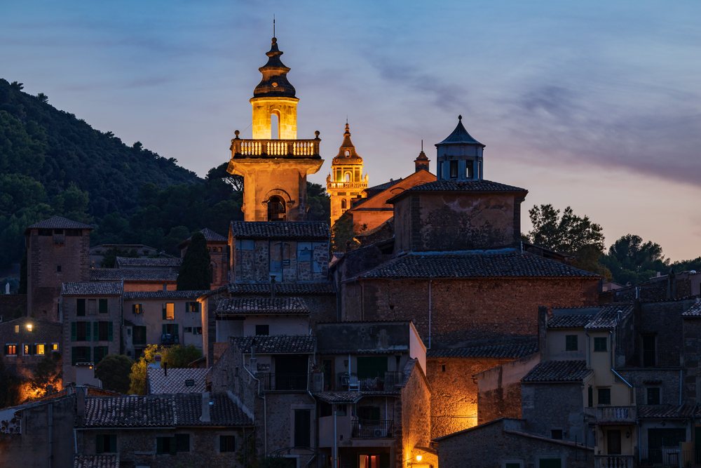 Skyline of Valdemossa at dusk with illuminated church towers and historic buildings below a twilight sky, Landscape, Lifestyle, Mallorca, Spain.