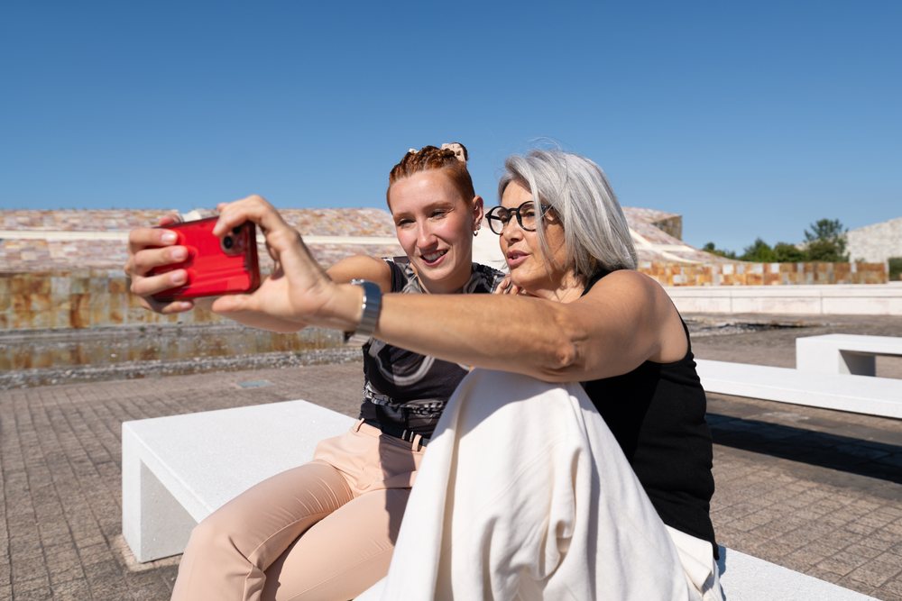 Two women sitting on a white bench outdoors take a selfie on a sunny day by modern architecture, businesswomen, Spain.