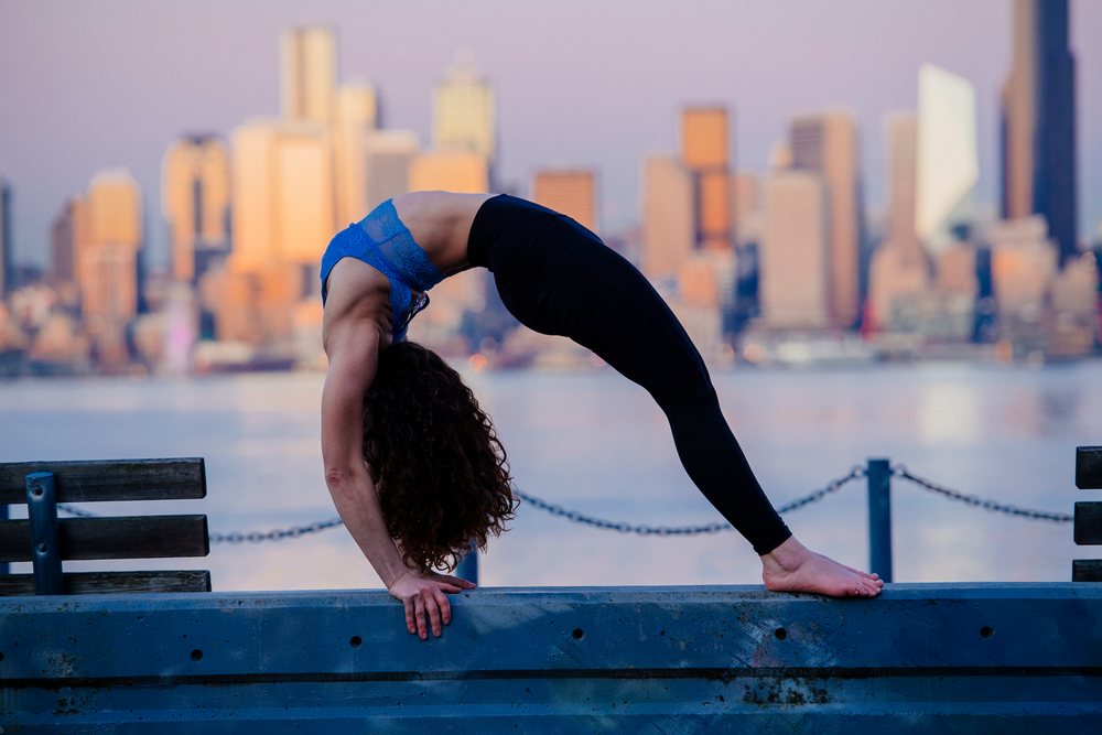 Woman performing a backbend yoga pose on a waterfront bench with Seattle city skyline at sunset, WA, USA.