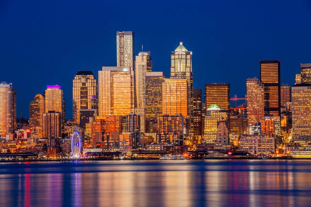 Nighttime panoramic view of Seattle waterfront city skyline with illuminated skyscrapers and reflections in ter under a dark blue sky, WA, USA.
