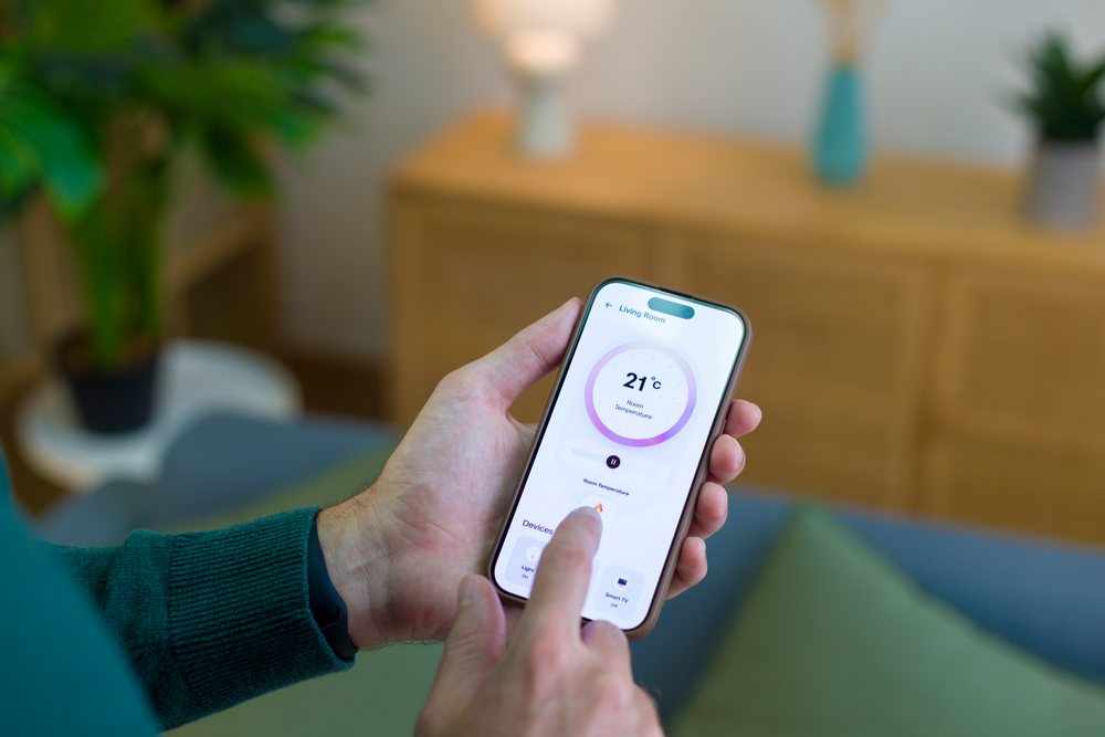 Person holding a smartphone displaying a temperature control app in a living room with a wood cabinet and indoor plant, technology, young adult, home.
