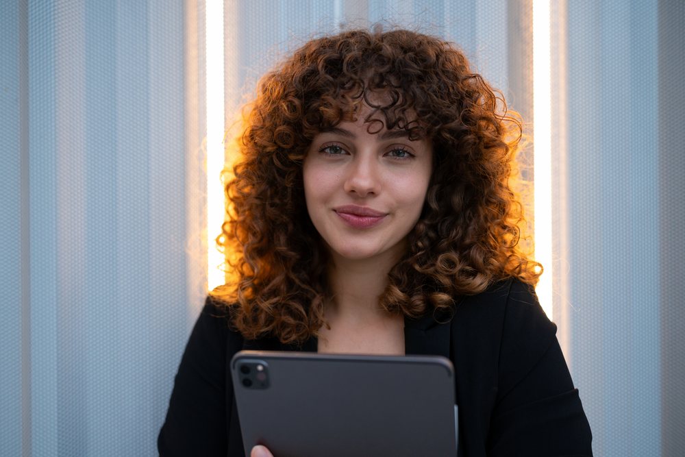 Young woman with curly hair holding a tablet computer and smiling in an indoor business setting with illuminated vertical panels, Madrid.