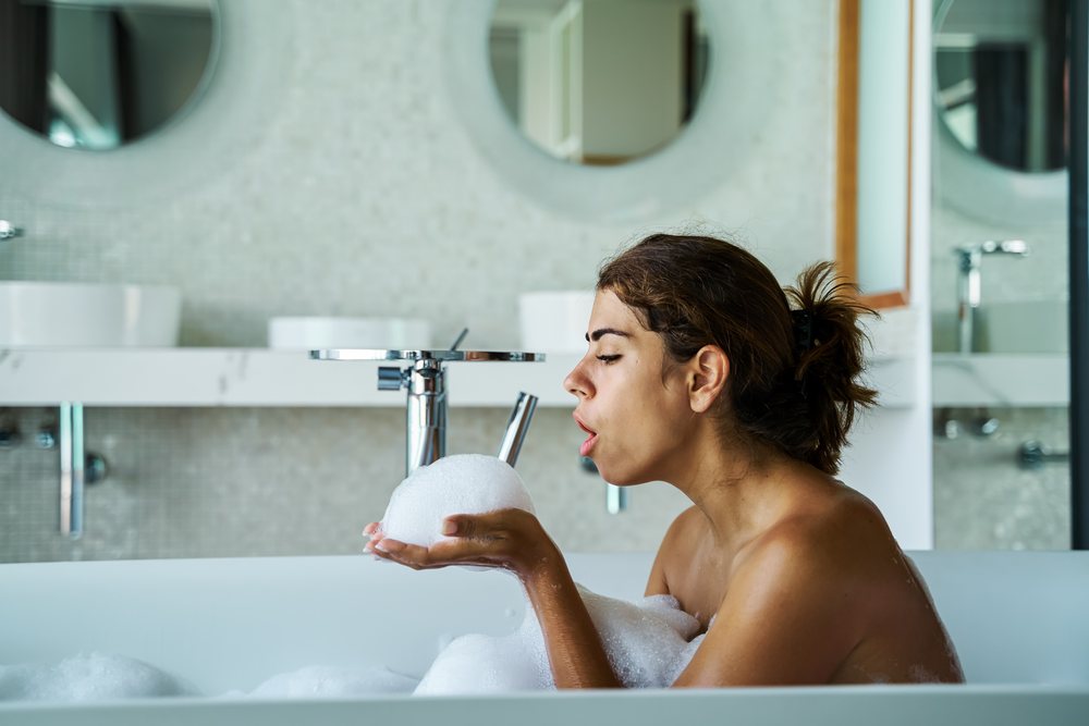 A woman blowing bubbles during a relaxing bubble bath in a modern bathroom, Maldives.