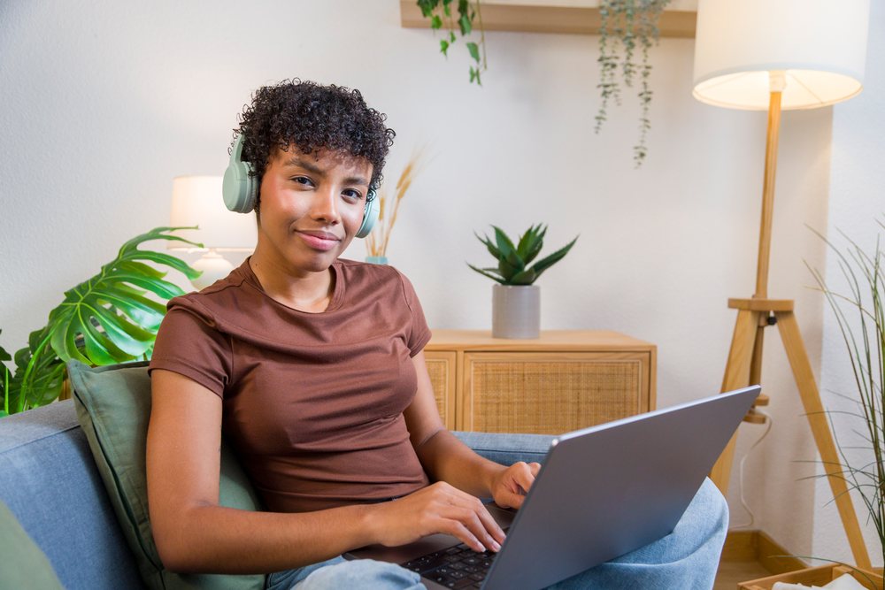 Woman with curly hair wearing headphones using a laptop while sitting on a sofa in a relaxed home setting.