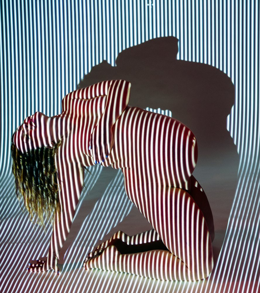 Pregnant woman kneeling nude with striped light projection and strong shadow in an indoor studio setting, pregnancy.