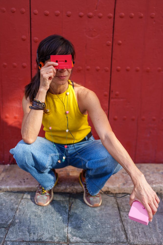 Woman squatting outdoors, listening to music with headphones connected to a pink cassette player and holding a red cassette tape, retro, vintage, Málaga, Spain.
