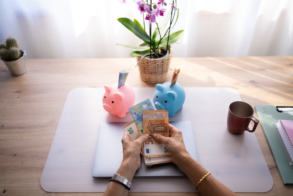 Hands counting euro banknotes on a wooden table with piggy banks, laptop, and plants in a home office setting, Spain.