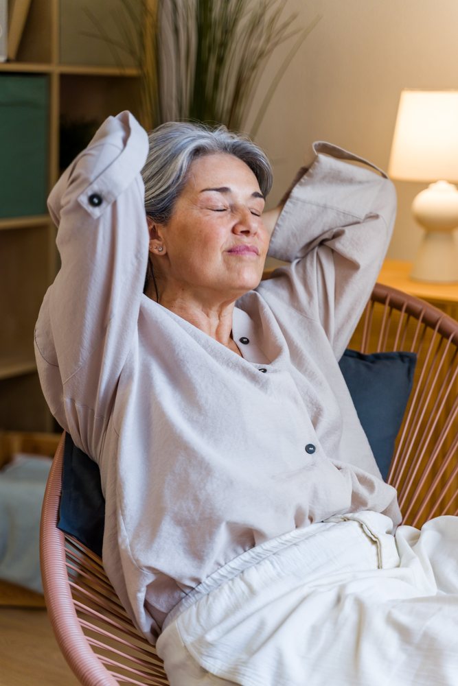 Gray-haired woman sits comfortably in a chair indoors with eyes closed and arms raised behind her head, relaxing peacefully, Spain.